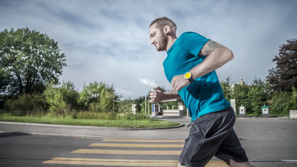 Caucasian man jogging on a city street crosswalk wearing a blue t-shirt and yellow watch.